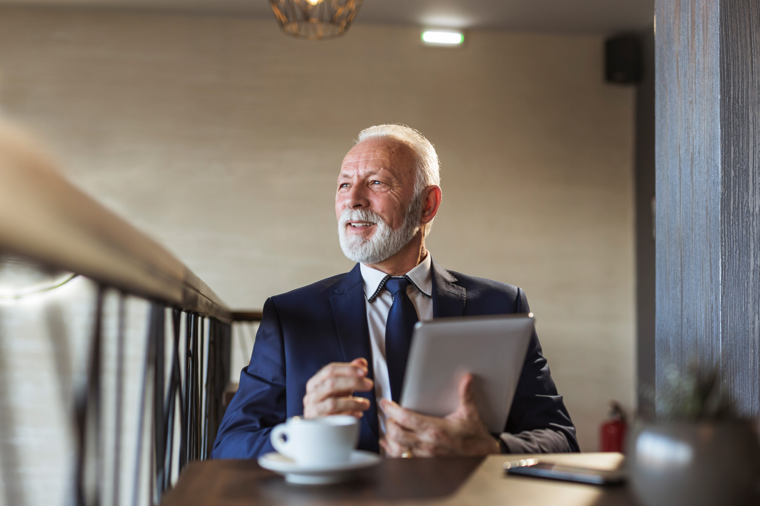 Senior businessman sitting at a restaurant table, drinking coffee and working on a tablet computer